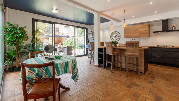 A dining area with a table and chairs, a kitchen with wooden cabinets, and a large window. - Villa 'L'acanthe' with Private Pool, Private Terrace, and Wi-Fi (Six-Fours-les-Plages)
