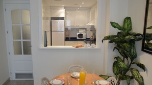 Un coin repas avec une table ronde dressée pour quatre personnes, une plante en pot et une vue sur la cuisine.