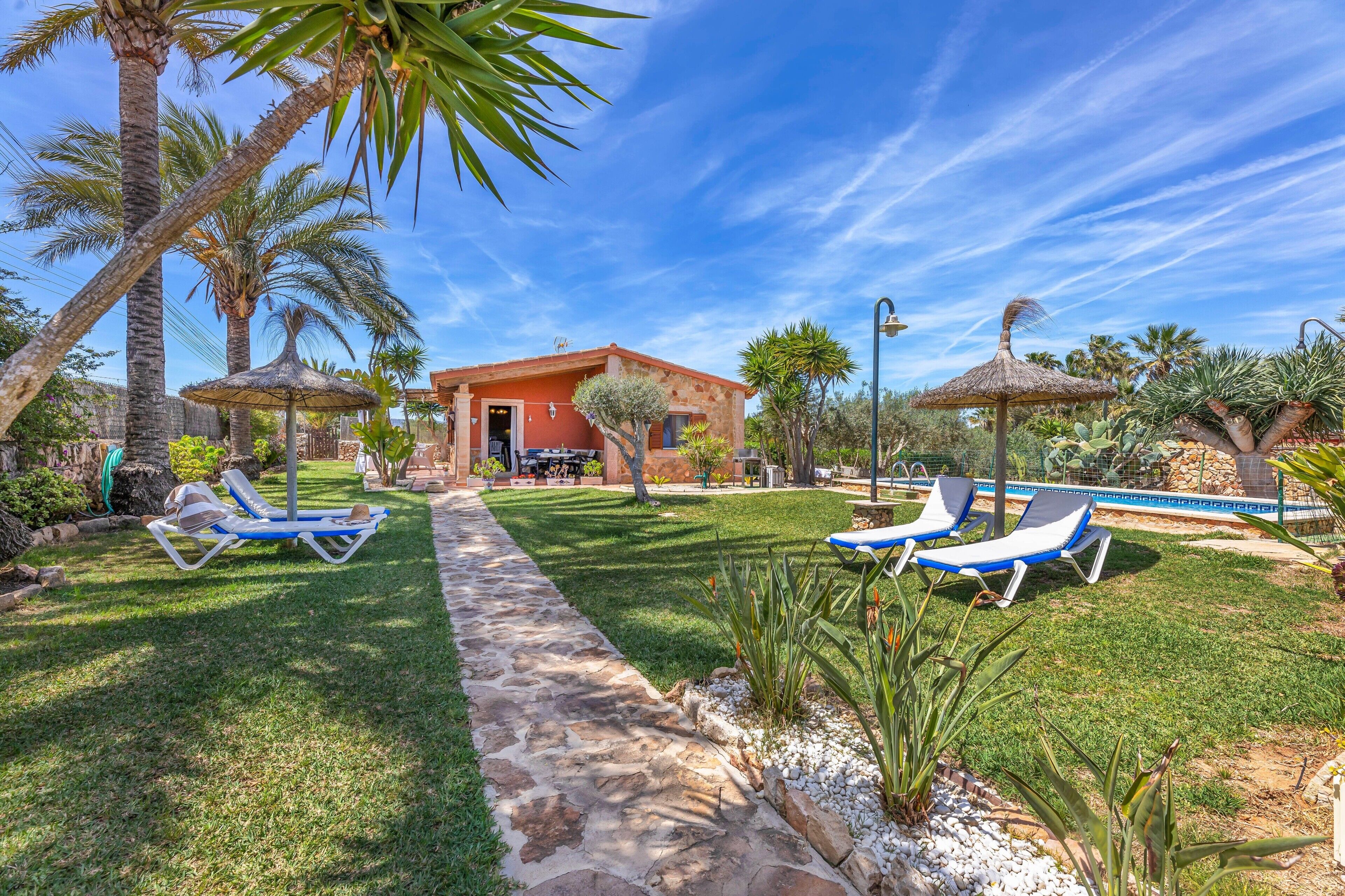 Un espace aménagé au bord de la piscine, avec des chaises longues, un parasol en chaume et un chemin de pierre menant à un bâtiment.