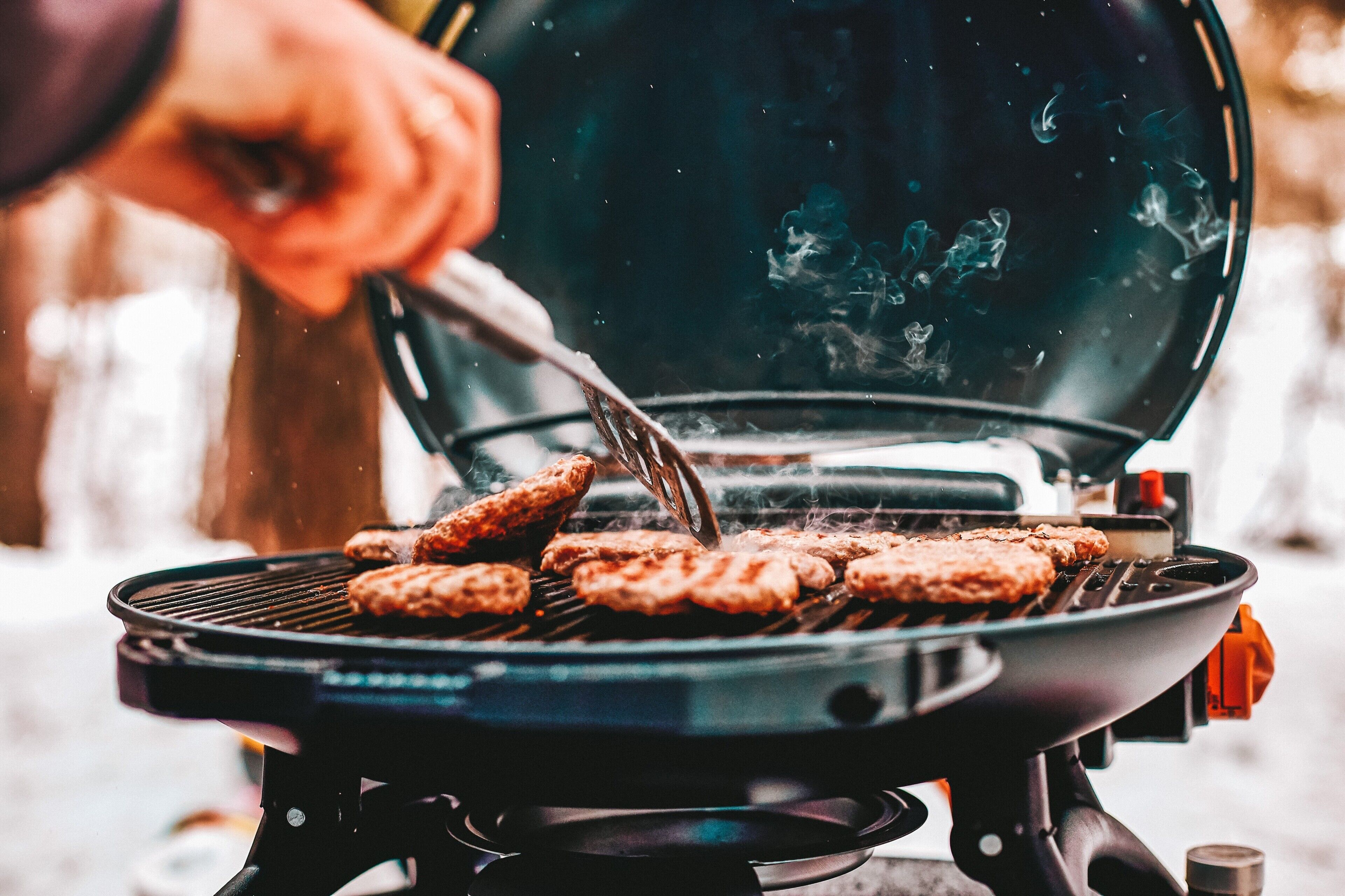 En grill med kött som tillagas, en person som använder en tång och en suddig utomhusbakgrund.
