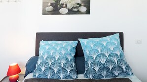 Una cama con dos almohadones estampados, una lámpara roja sobre una mesita de noche y un cuadro colgado en la pared encima de la cama.
