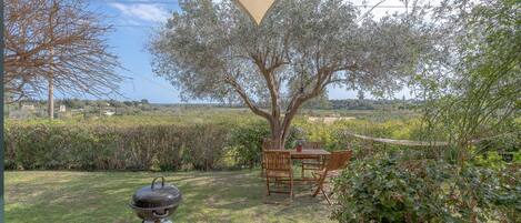 A backyard with a barbecue grill, a wooden table and chairs, and a large tree providing shade.