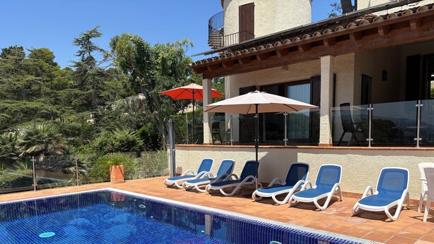 A swimming pool with blue mosaic tiles, surrounded by lounge chairs and umbrellas, with a building in the background.