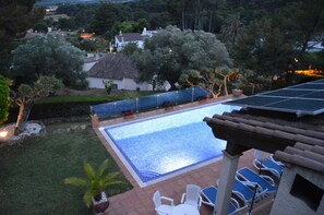 A swimming pool with a tiled deck, surrounded by a garden with various plants and a white building in the background. - Villa 'Estrella' with Mountain View, Wi-Fi and Air Conditioning (La Torre Vella)
