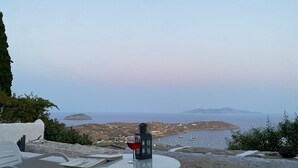 Une table avec un verre de vin rouge, un livre et une lanterne, sur un balcon donnant sur un paysage côtier avec des bateaux et des îles au loin.