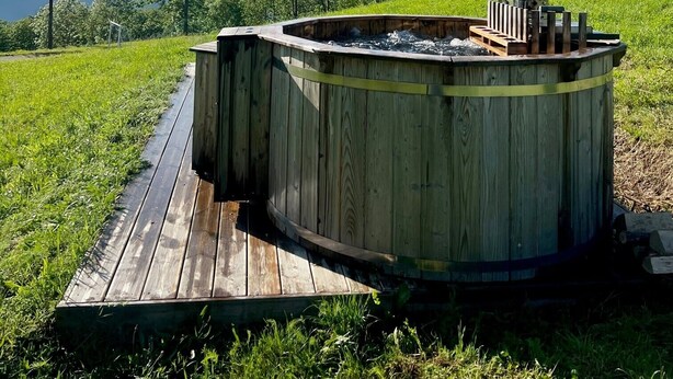 Puinen poreallas, jossa on metallinen savupiippu, ja sen ympärillä puinen terassi. Taustalla kohoavat vihreät kukkulat ja kirkas taivas.