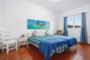 Una camera da letto con un letto, una sedia, un comodino, un quadro raffigurante una scena tropicale e una finestra con tende.
