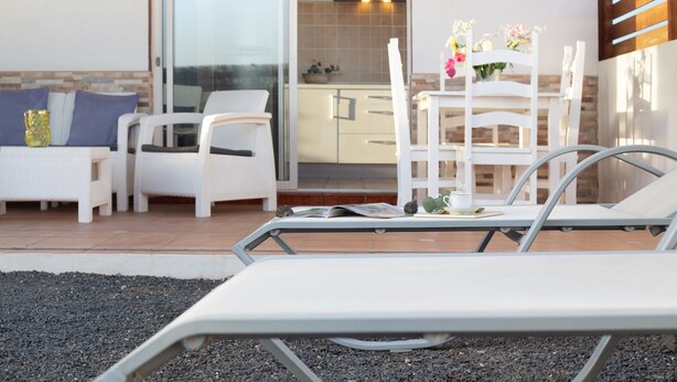 Un balcón con mobiliario blanco, una mesa con un libro y una silla con un florero.