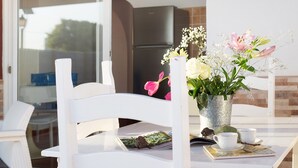 Una silla blanca, una mesa con un florero y un refrigerador al fondo.