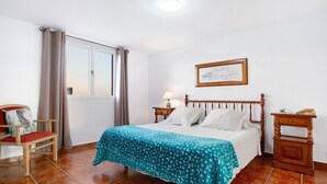 Un dormitorio con una cama, una silla, una mesita de noche, una lámpara, una ventana y un cuadro enmarcado en la pared.