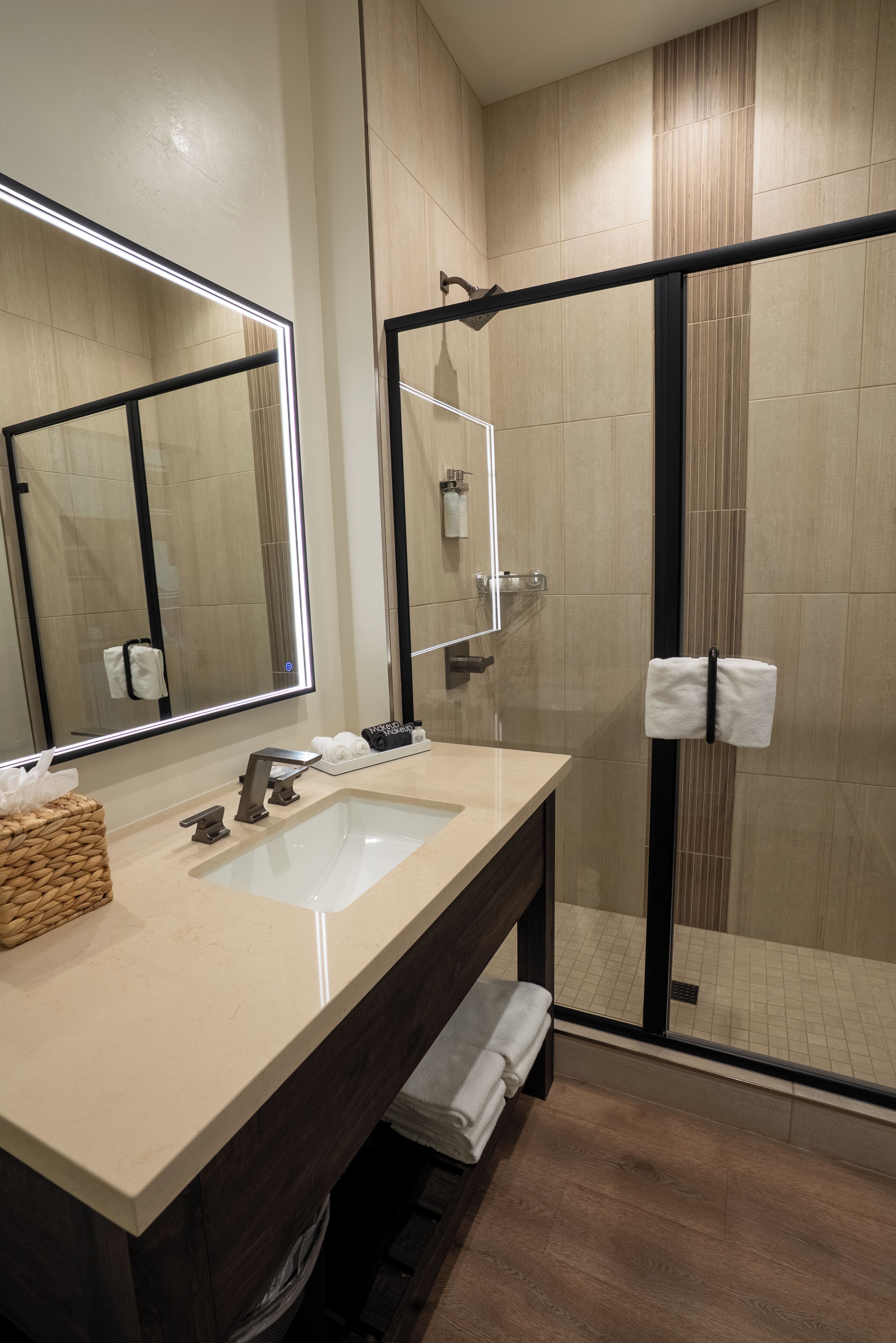 A modern bathroom with a large mirror, dual sinks, a glass shower enclosure, and a wooden floor.