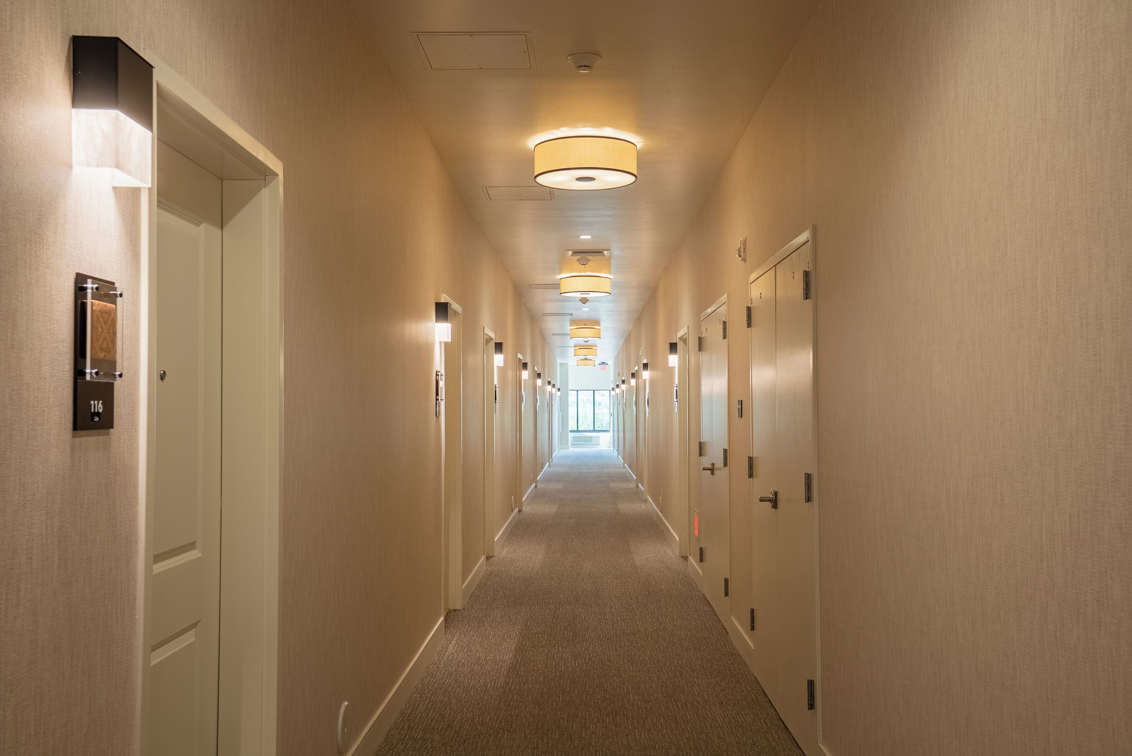 A hotel hallway with doors on both sides, overhead lighting, and a carpeted floor.