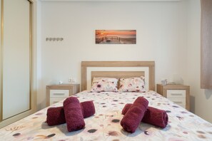A neatly made bed with floral bedding, two nightstands with lamps, a wooden headboard, and a sunset painting on the wall.