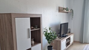 A modern living room with a wooden cabinet, a television, a small table with a plant, and a shelf with decorative items. - Apartment 'Carpediem in Spanje' with Lake View, Shared Pool and Wi-Fi (San Miguel de Salinas)
