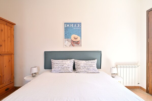 Une chambre à coucher avec un lit, deux lampes de chevet, une armoire en bois, une affiche accrochée au mur et un radiateur.
