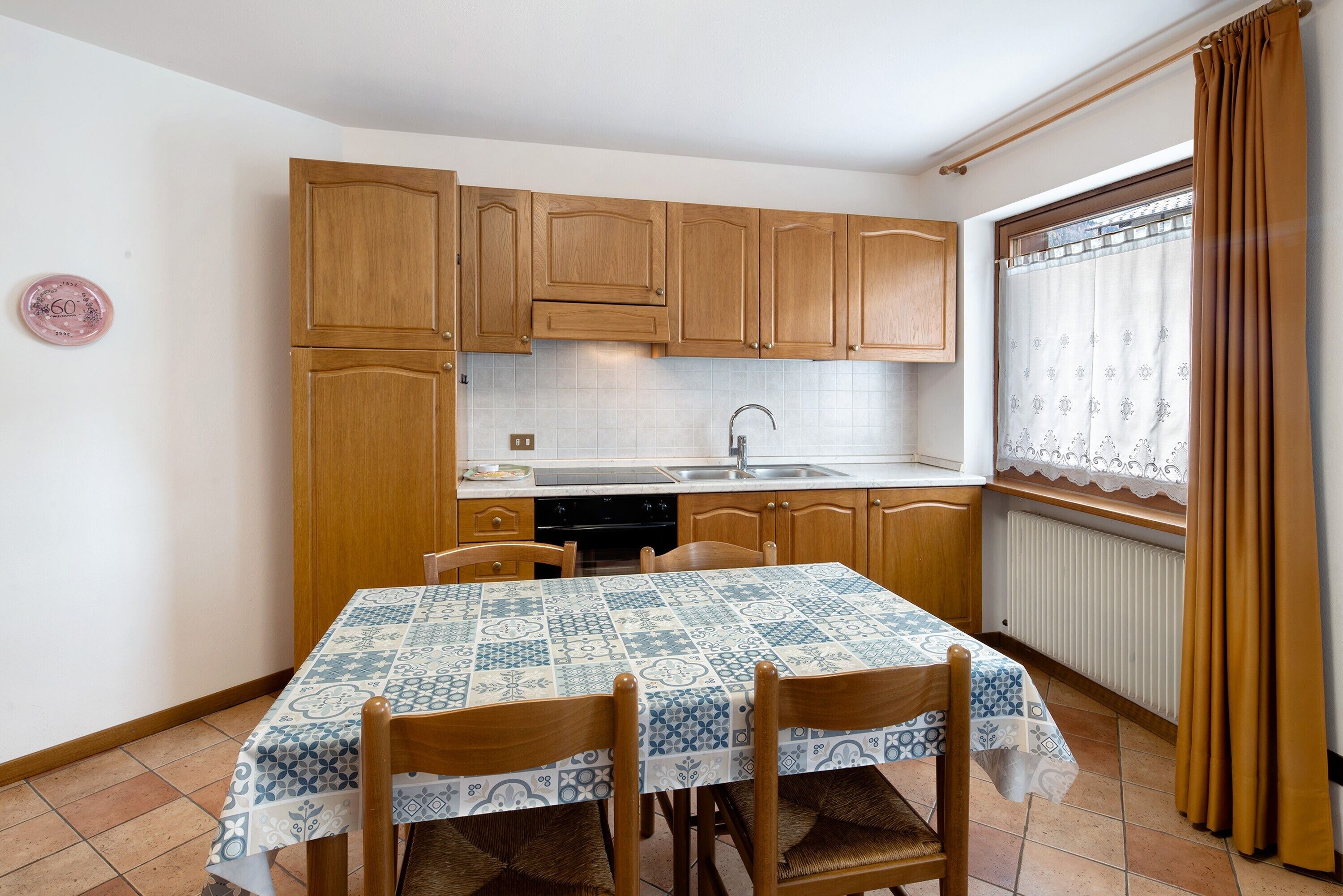 A kitchen with wooden cabinets, a tiled backsplash, a dining table with chairs, and a window with curtains.