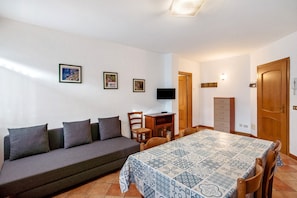 A room with a sofa, a dining table with a patterned tablecloth, a TV on a stand, and a wooden cabinet.