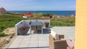 A patio with a table and chairs, a potted plant, and a view of the sea and houses in the background. - Lovely home in Hjørring with sauna (Hjørring)
