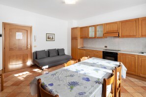 Une cuisine compacte avec des meubles en bois, une table à manger recouverte d’une nappe à motifs floraux, un canapé et une porte sur laquelle est affiché un avis.