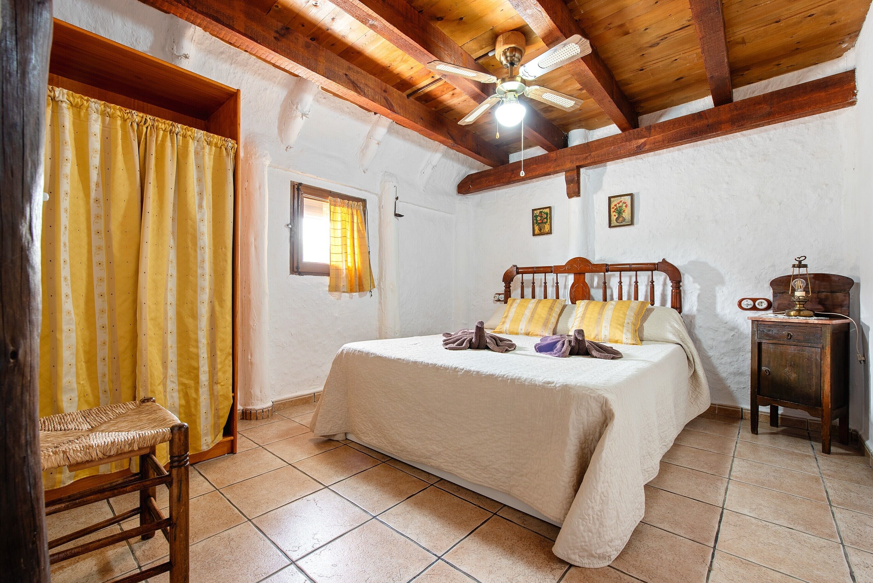 Une chambre avec un plafond en bois, un lit recouvert d’une courtepointe blanche, une table de chevet en bois et une fenêtre avec des rideaux.