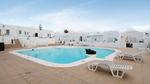 Una piscina con sillones de descanso, una palmera y edificios blancos.