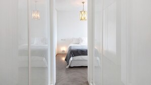Un pasillo estrecho que conduce a un dormitorio con una cama, dos luces fijadas a la pared y una puerta blanca.
