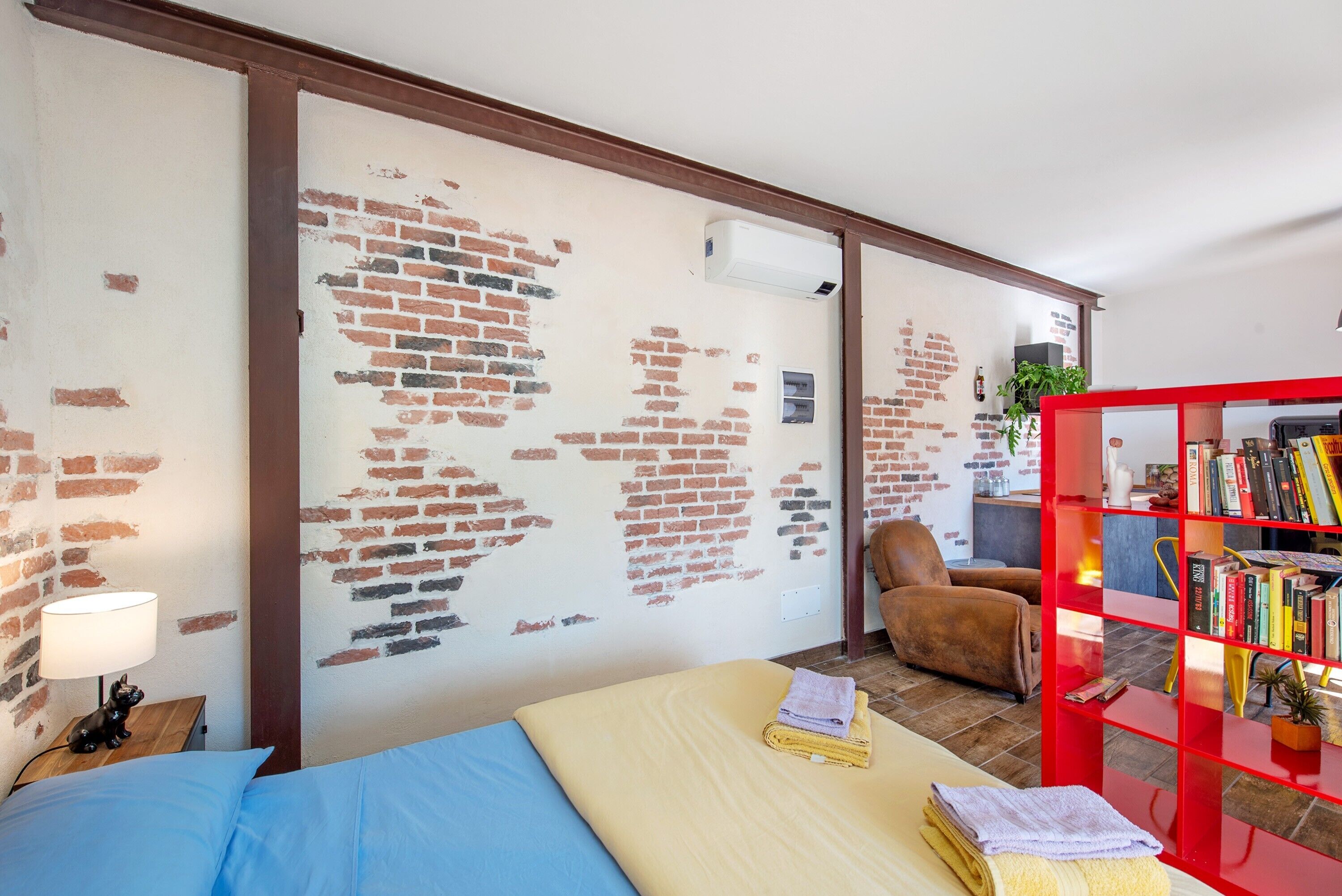 Ein Zimmer mit Backsteinwand, einem Bett mit blau-beiger Bettwäsche, einem roten Bücherregal und einem braunen Ledersessel.