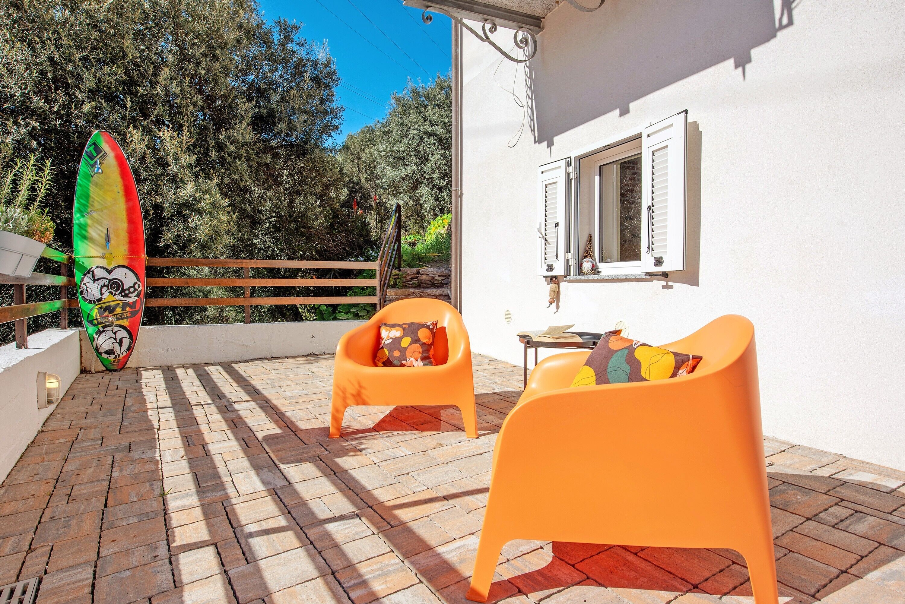 A patio with two orange chairs, a surfboard, and a white building with shutters.