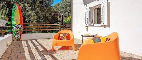 A patio with two orange chairs, a surfboard, and a white building with shutters.