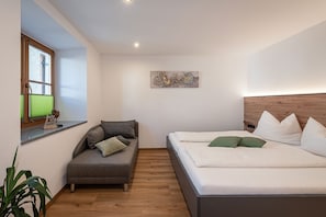 A bedroom with a bed, a sofa, a window with blinds, a wall-mounted artwork, and a plant.