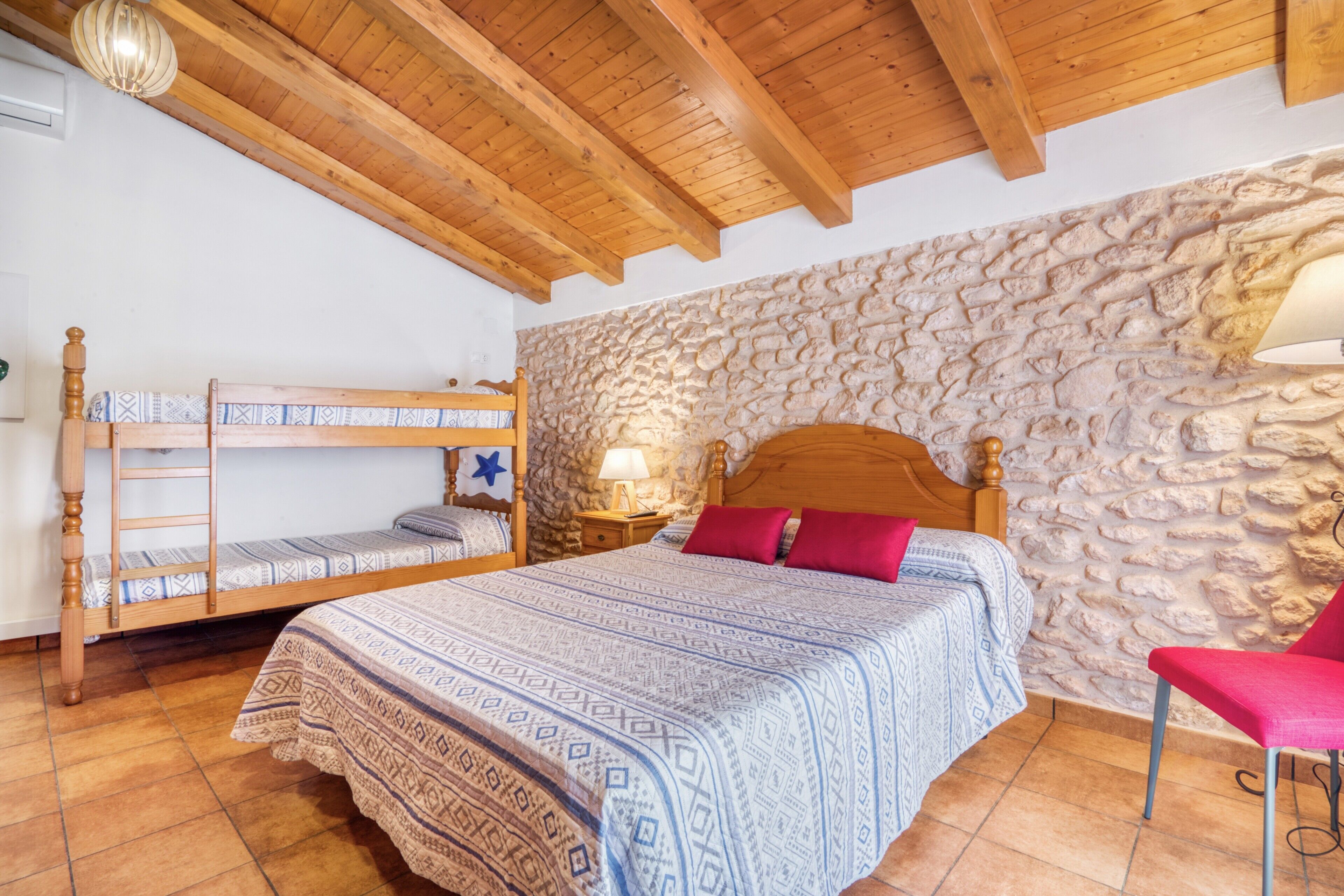 A bedroom with a bunk bed, a single bed with patterned bedding, a stone wall, and a wooden ceiling.