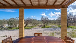 A covered outdoor seating area with a wooden table and chairs, a pergola structure, and a view of a landscaped garden with trees and a gravel path. - Holiday Home 'Casa Degli Ulivi' with Private Terrace and Wi-Fi (Marzamemi)
