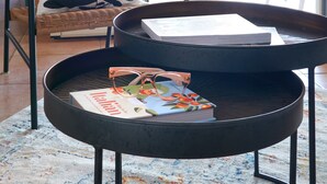 Une chaise en osier avec un coussin noir et blanc à motifs de feuilles, une table ronde en métal sur laquelle se trouvent un livre et des lunettes, et une plante en pot à l’arrière-plan.