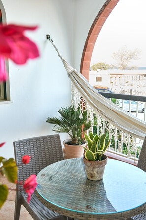 Un balcón con una silla de mimbre, una mesa de vidrio, plantas en macetas y una hamaca.
