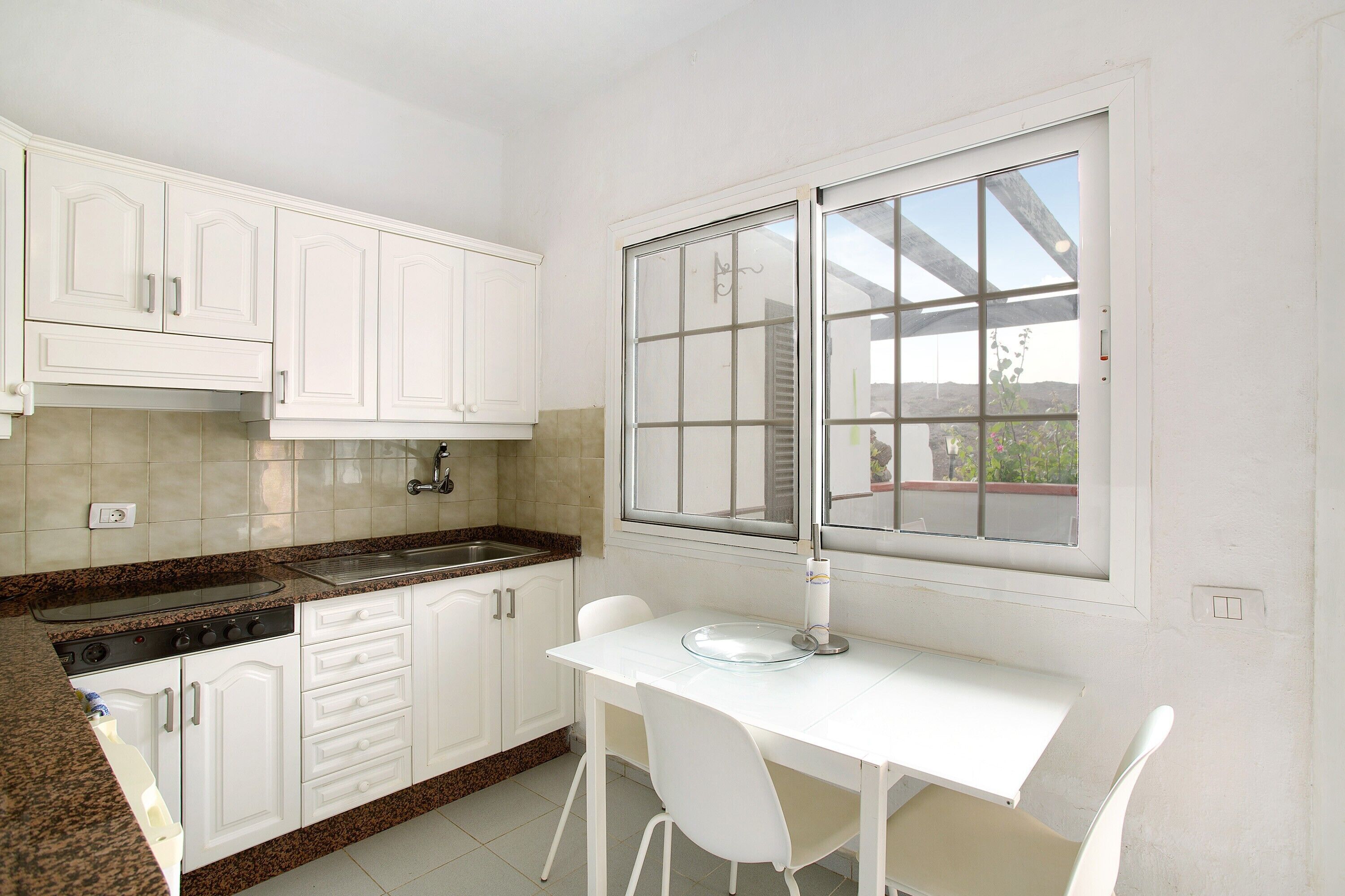 Una cocina con armarios blancos, encimera de granito, ventana con vistas y una mesa de comedor blanca con sillas.