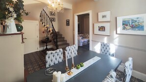 Un comedor con una mesa oscura, velas blancas y sillas estampadas. Una escalera con barandilla negra conduce a otra habitación.
