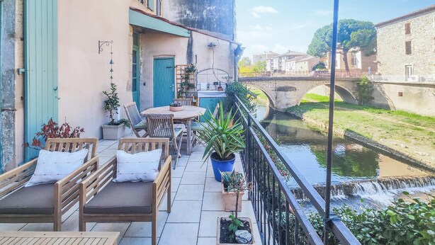 Un balcón con mesa y sillas, un pequeño jardín y vistas a un canal con un puente.
