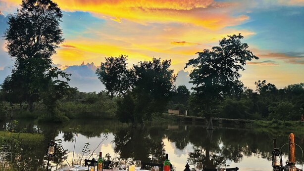 An outdoor dining setup with a table set for two, surrounded by chairs, near a body of water with trees in the background. - Bandhavgarh Meadows (Manpur)