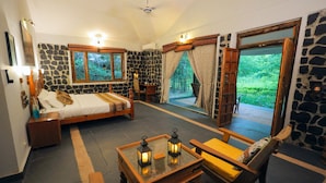 A bedroom with a stone wall, a bed, a nightstand, a wooden table with lanterns, and a view of greenery through a window. - Bandhavgarh Meadows (Manpur)