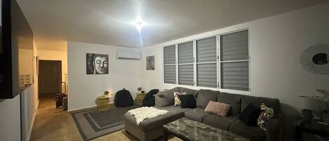 A living room with a grey sofa, a glass coffee table, and a small rug. There are two side tables with decorative items, a wall-mounted air conditioner, and a window with blinds.