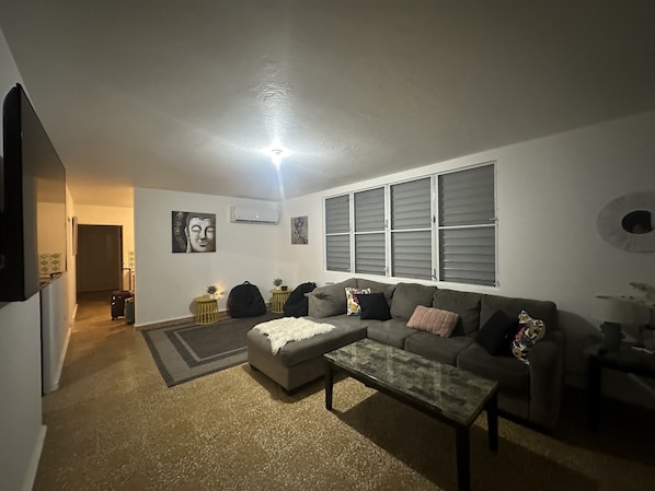 Un salón con un sofá gris, una mesa de centro de vidrio y una alfombra pequeña. Hay dos mesitas de noche con adornos, un aire acondicionado de pared y una ventana con persianas.