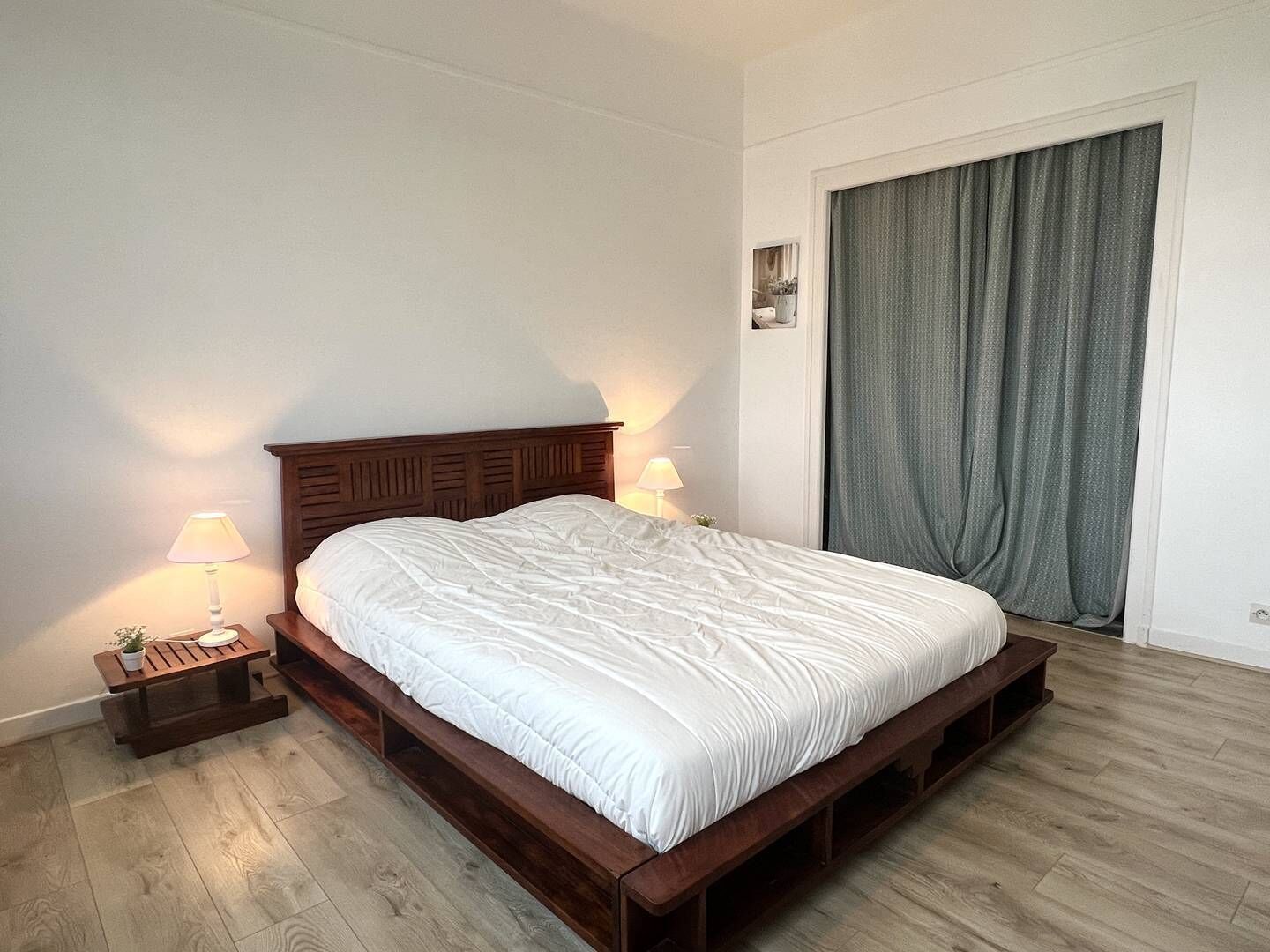 Camera da letto con un letto in legno, due lampade sui comodini, un piccolo tavolino da notte con una pianta e un'ampia finestra con tende.