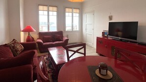 A living room with a red sofa, a red table, a TV on a red cabinet, and a lamp. - 4-person apartment near the beach in Sables-d’Or-les-Pins | Ref 107 (FREHEL)