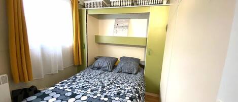 A compact bedroom with a bed, a window with curtains, a radiator, and a small shelf with items.