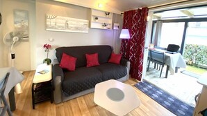A modern living room with a dark grey sofa, red cushions, a white coffee table, and a small side table. There is a painting on the wall, a lamp, and a view of the outside through a large window. - Charming chalet located in a 4-star  campsite - 6 people (GUIDEL-PLAGES)