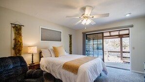 A bedroom with a bed, a chair, a nightstand, a lamp, a ceiling fan, and a large window. - Mingus Mountain Vista: Views, Shopping, Relaxation. 20 Minutes to Sedona  (Cottonwood)