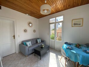 Una habitación con un sofá gris, una pequeña mesa de centro, una mesa de comedor preparada para dos personas y una ventana con vistas al exterior.