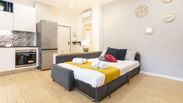 Una habitación de hotel moderna con kitchenette, una cama con manta amarilla y un sofá gris.