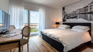Quarto de hotel com cama, escrivaninha, cadeira, televisão e uma janela grande com cortinas.
