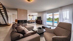 Uma sala de estar moderna com um conjunto de sofá, uma mesa de centro e uma televisão. Uma escada leva a outro nível, e uma piscina é visível pelas portas de vidro.